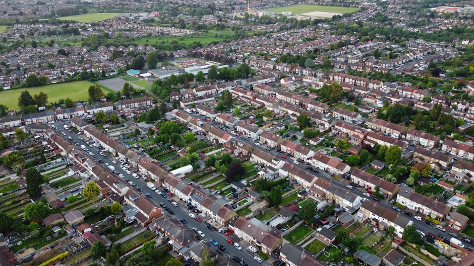 An aerial view of a residential area featuring neatly aligned terraced houses with small front gardens and paved driveways. The streets are lined with parked cars, and the gardens display well-maintai