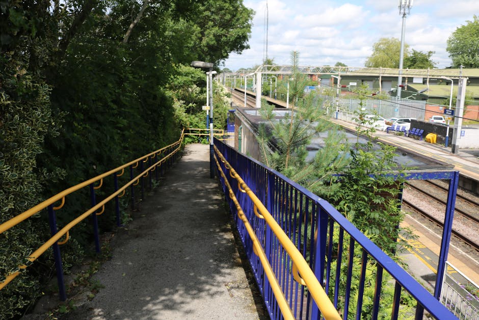 A narrow pathway along the station forecourt gardens features a textured asphalt surface with shadows cast by nearby trees. On the left side, dense green foliage and shrubs grow close to the path, pre